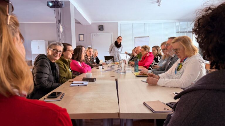 Długi stół przy którym siedzą uczestniczki spotkania. Na końcu stoi prowadząca, trzyma ręce za plecami. W tle biała tablica.